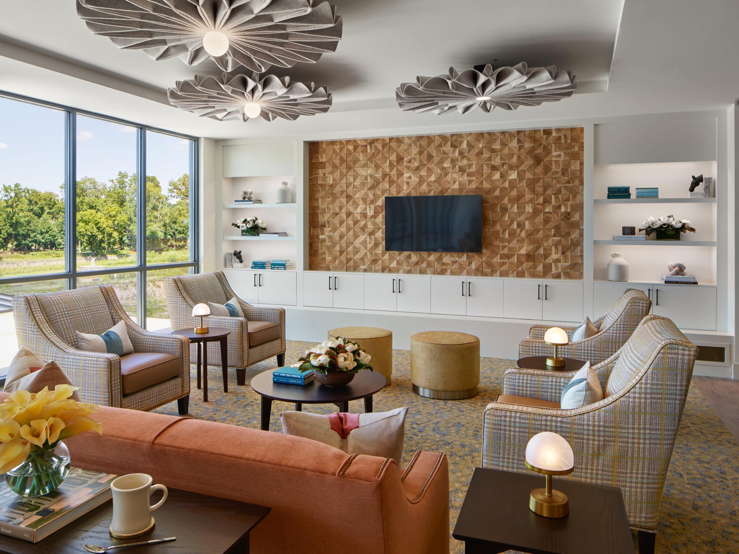 Stylish living room features modern decor, cozy seating, geometric lighting, and expansive windows overlooking greenery. Warm tones and unique textures enhance the inviting ambiance.