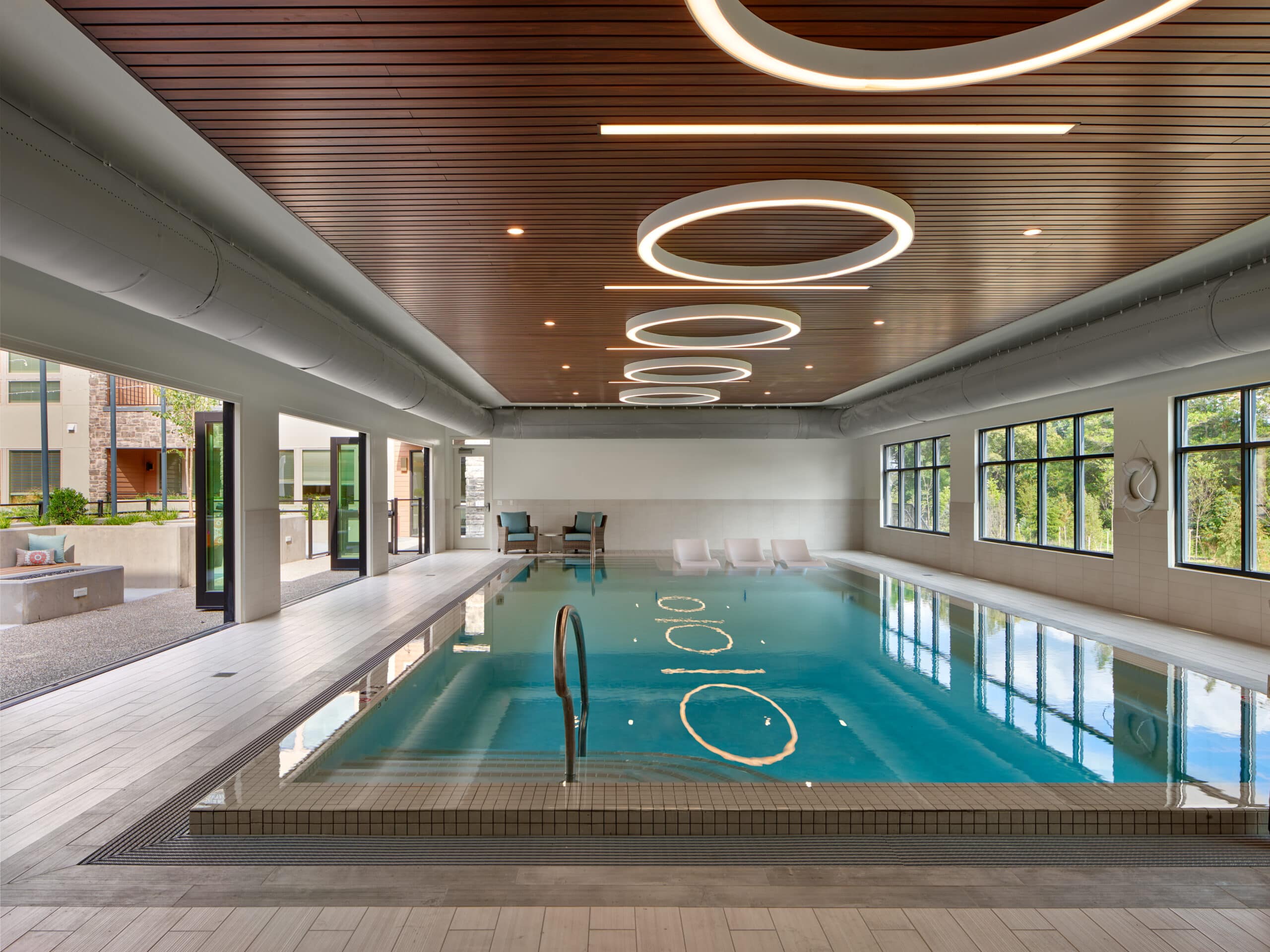 Indoor pool with modern lighting and lounge chairs. Large windows provide natural light. Open doors connect to an outdoor seating area.